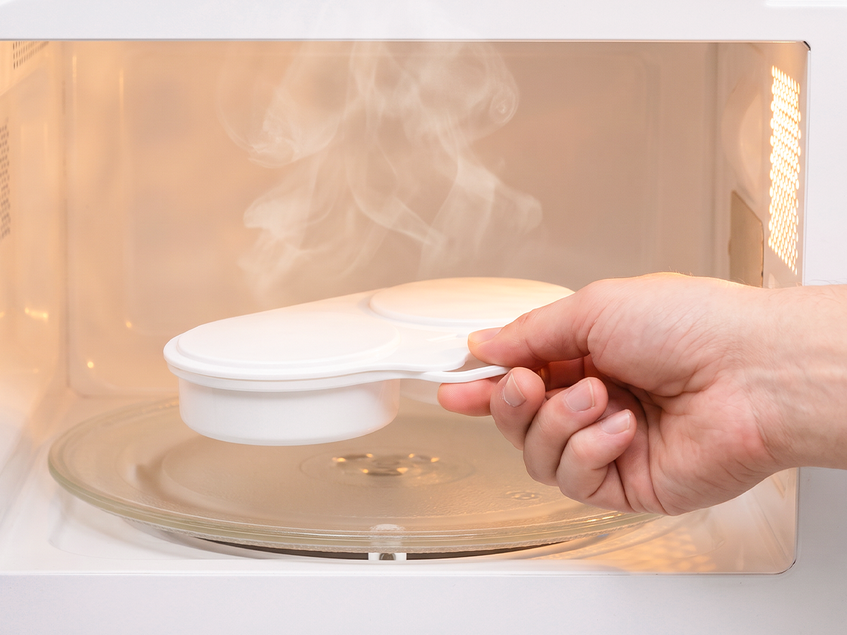 White dual-cavity egg steamer held by its handle inside a microwave