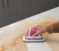 A pink and white flat scrubber moves across a crumb-covered white countertop.